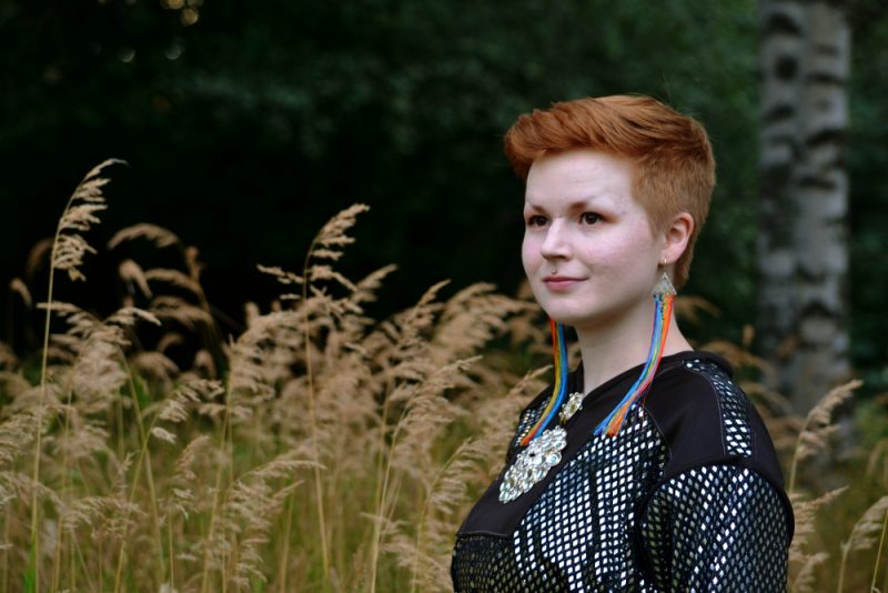 Kuvassa on Anne Olli, joka seisoo pitkien heinien edessä saamenpuvussa ja sateenkaarikorvakoruissa.