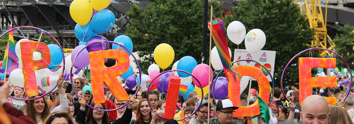 Kuva Pride-tapahtumasta. Värikäs ihmisjoukko marssimassa sateenkaarilippuja mukanaan, osa ihmisistä pitelee hulavanteisiin kiinnitettyjä suuria, oranssi-pinkkejä kirjaimia, jotka tavaavat sanan "PRIDE". Taustalla ilmapalloja ja puita.