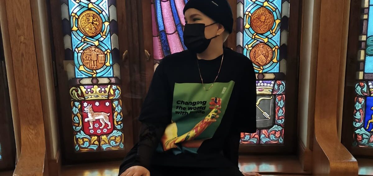 Maskiin ja pipoon pukeutunut Piisu Tuominen istuu koristellisen ikkunan edessä ja katsoo sivulleen.