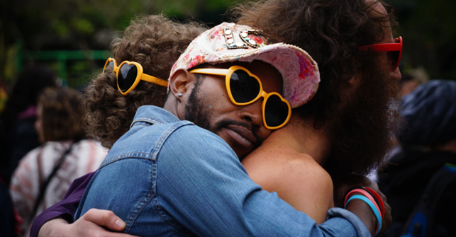 Tummaihoinen parrakas mies lippalakissa ja sydänaurinkolaseissa halaa toista henkilöä takaapäin.