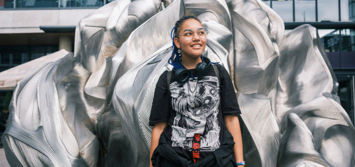 Sateenkaarinuori Julia seisoo Helsingin Itäkeskuksen Tallinnanaukiolla metallisen veistoksen edessä. Hänellä on musta printti t-paita ja mustat housut sekä rastat ja reppu selässä. Julia on tummaihoinen.