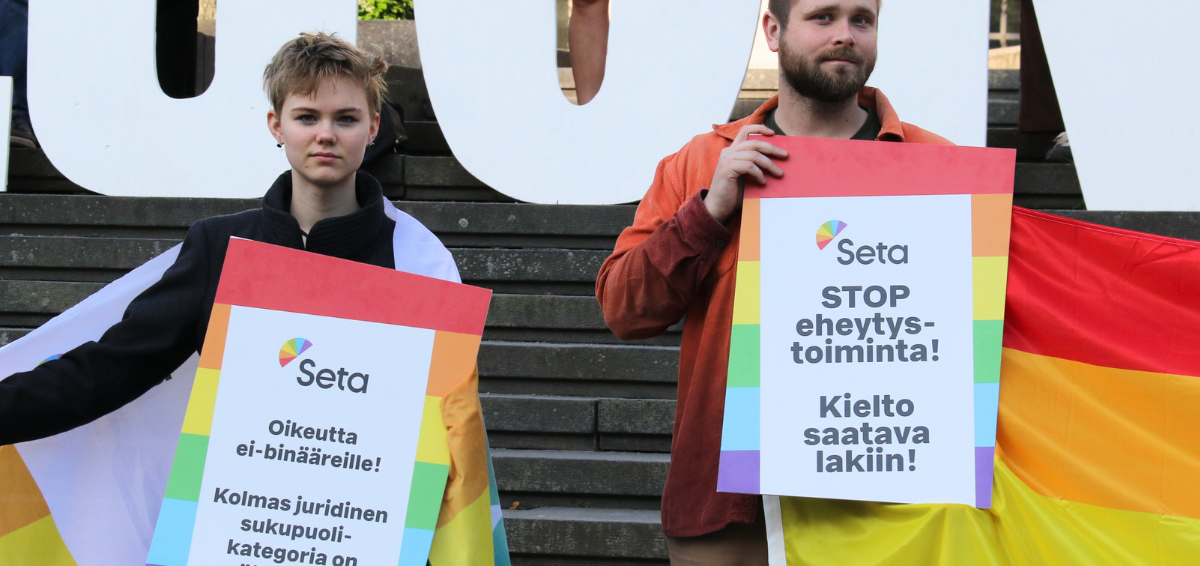 Sinitukkainen ja vakava sateenkaarinuori ja Setan hallituksen jäsen, parrakas lyhythiuksinen mieshenkilö pitävät mielenosoituskylttejä Säätytalon edessä kevät-kesällä 2023. Molemmilla on myös sateenkaariliput, joita he pitelevät.