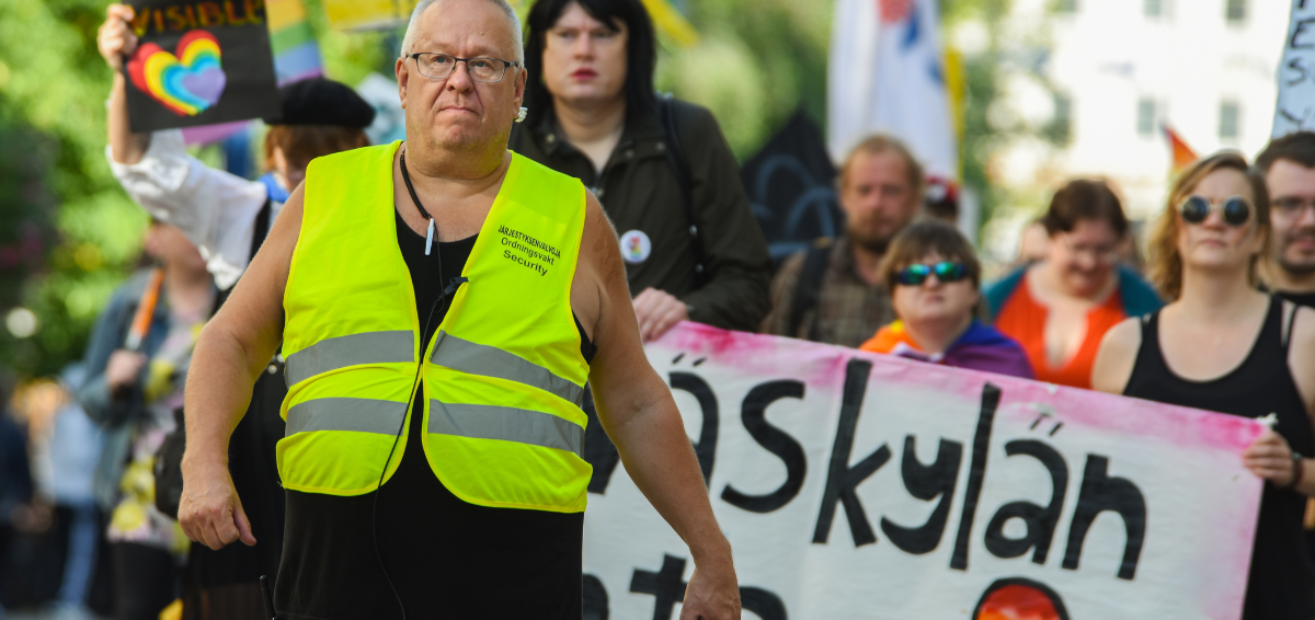 Jyväskylä Pride -kulkue kuvattuna edestä. Ensimmäisenä marssii keski-ikäinen valkoihoinen silmälasipäinen Petri Janhunen huomioliivi päällään. Taustalla kannetaan kylttejä. Ilmeet ovat vakavia.