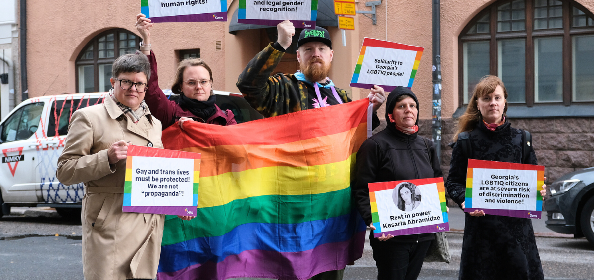 mielenosoittajia sateenkaarilipun ja kylttien kanssa, etualalla on kynttilöitä maassa, taustalla näkyy georgian lippu ja lähetystön ovi