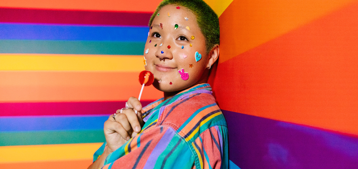 Nuori henkilö lyhyillä, ajetuilla hiuksilla katsoo ylös kameran yläpuolelle ja hymyilee nojaten sateenkaaren väriseen seinään. Hän on pukeutunut värikkääseen raitapaitaan ja pitää kädessään tikkaria. Kasvoilla hänelle on liimattuja monia tarroja ja jalokiviä. Nenässään hänellä on septum-lävistys.