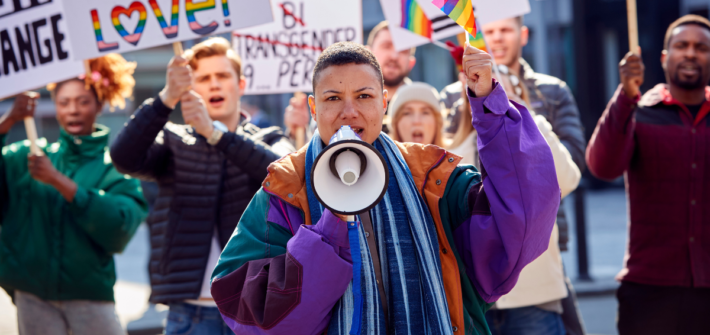Etualalla henkilö Pride-kulkueen kärjessä huutamassa megafoniin. Takana ihmisiä kylttien kanssa.