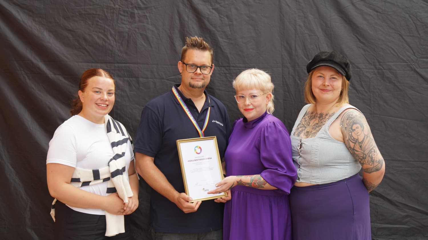 Neljä henkilöä seisoo ulkona ison telttakankaan edessä. Keskellä oleva silmälasipäinen mies pitää käsissään diplomia.