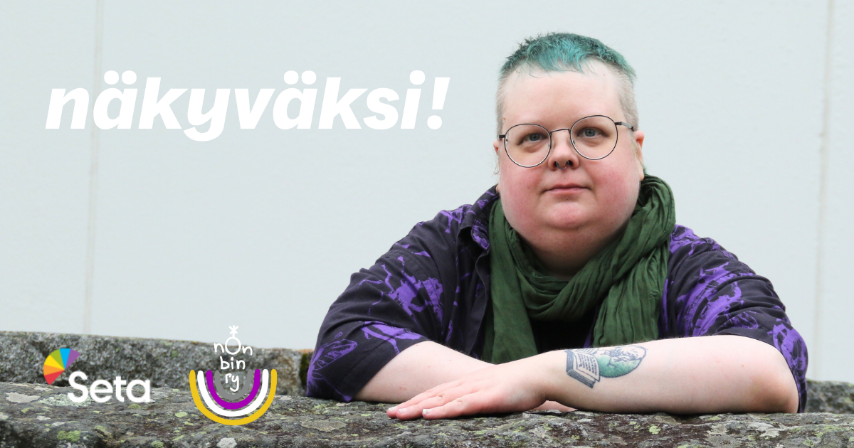 Henkilöllä on vihreä lyhyt tukka, silmälasit, vihreä huivi ja violettimusta paita. Kädessä on tatuointi. Hän seisoo kiviaidan takana ja on ristinyt kädet kiviaidan päälle. Kasvoilla on päättäväinen ja vakava ilme.