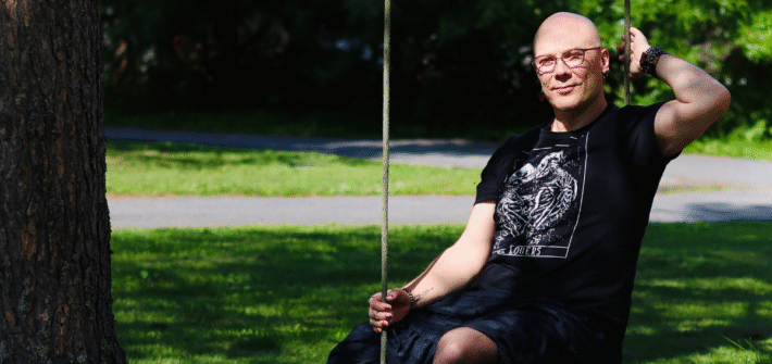 Mustiin pukeutunut kalju ja silmälasipäinen henkilö keinuu narukeinussa.