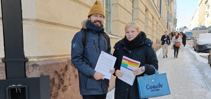 Ulkona monikerroksisen talon edessä seisoo kadulla kaksi henkilöä talvivaatteissa. He pitelevät asiakirjoja.