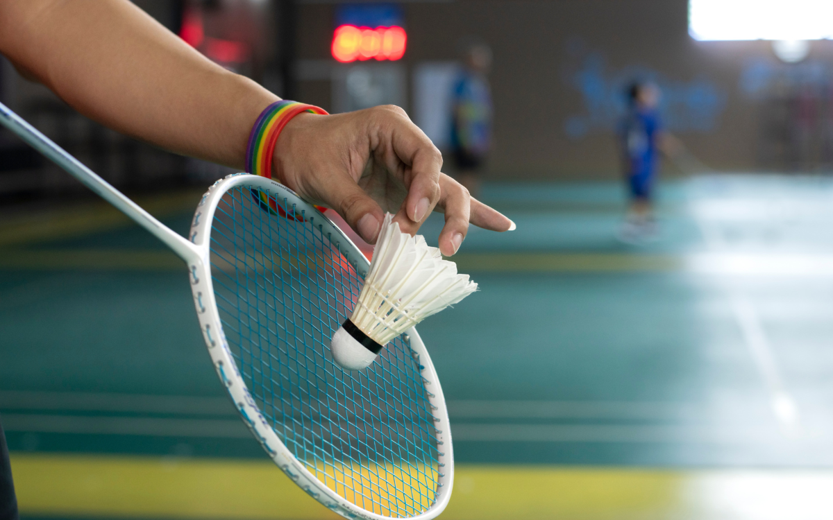 Käsi pitelee sulkapalloa sulkapallomailaa vasten liikuntahallissa. Ranteessa on sateenkaarinauha.