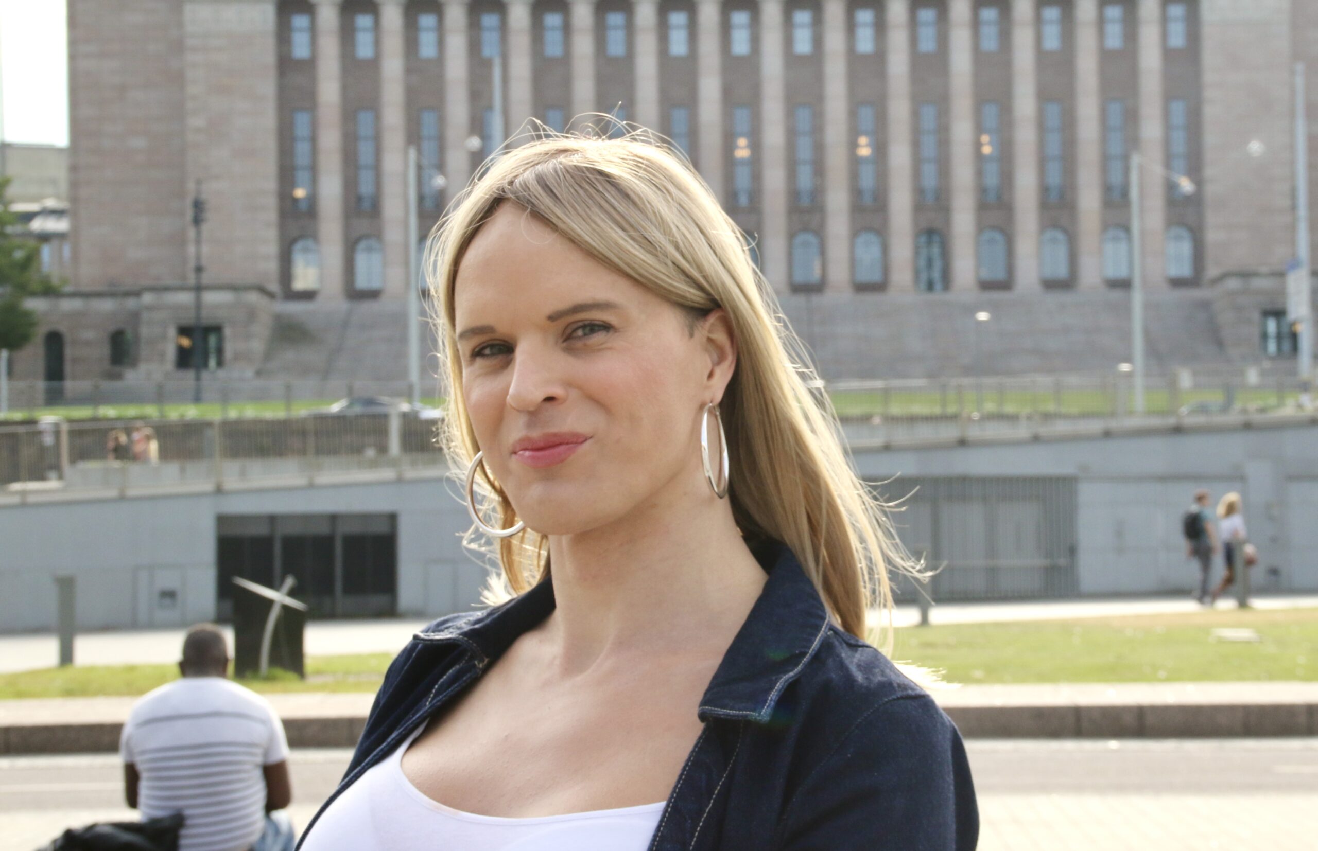 Henkilöllä on vaalea pitkä tukka, hopeiset korvarenkaat ja tumma farkkutakki. Hänen taustallaan näkyy Helsingin eduskuntatalo. Kasvoilla on hymy.